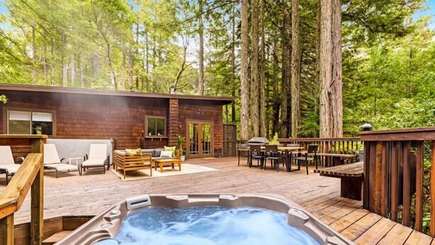 Una cabaña de madera con jacuzzi y zona de estar exterior rodeada de árboles altos.