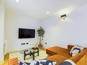 Ein modernes Wohnzimmer mit einem an der Wand montierten Flachbildfernseher, einem Glastisch, einem Blumentopf und einem braunen Sofa mit gemusterten Kissen.