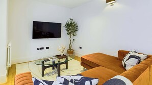 A modern living room with a flat-screen TV mounted on the wall, a glass coffee table, a potted plant, and a brown sofa with patterned cushions. - Light-filled Maida Vale Mews Retreat (Greater London)