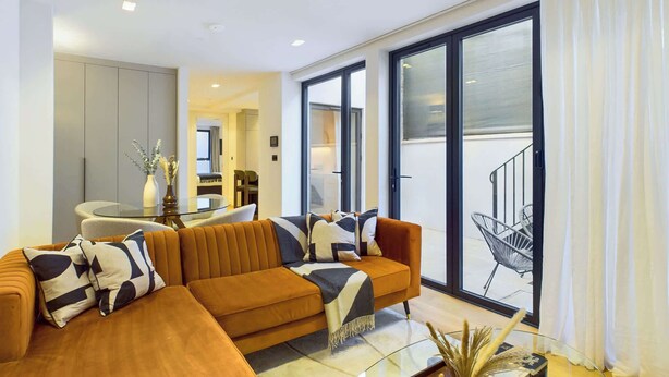 A modern living room with a large sectional sofa, a glass coffee table, and a view of a balcony with a staircase. - Light-filled Maida Vale Mews Retreat (Greater London)