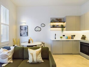 Ein modernes Wohnzimmer mit einem grünen Sofa, einem kleinen runden Tisch und einer Küchenzeile mit Einbauofen und Kaffeemaschine.