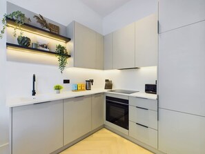 A modern kitchen with white cabinets, a black stove, and a built-in oven. There are shelves with decorative items and a hanging plant.