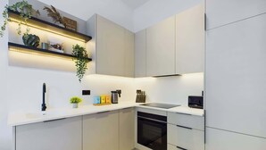 Une cuisine moderne avec des armoires blanches, une cuisinière noire et un four encastré. On y trouve des étagères ornées d’objets décoratifs et une plante suspendue.