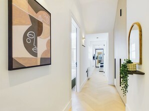 A modern hallway with a minimalist design, featuring a geometric abstract painting on the left, a sleek white door, and a plant on a console table.