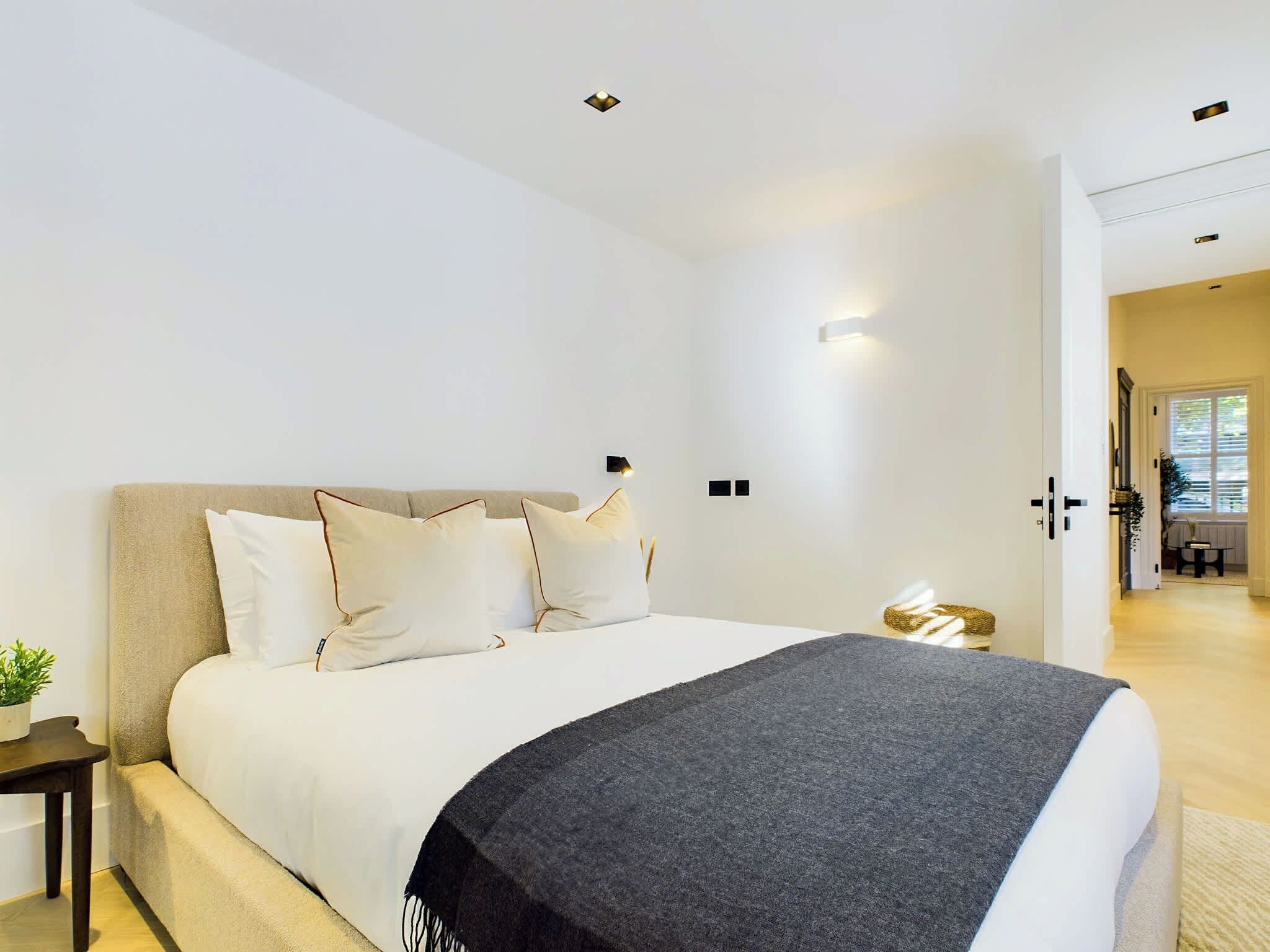 A modern hotel room with a large bed, a bedside table with a plant, and a view of the hallway.