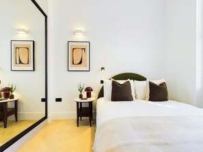 A hotel room with a large bed, a round side table with a lamp, a small table with a plant, and two framed abstract paintings on the wall. - Modern Maida Vale Apartment (Greater London)