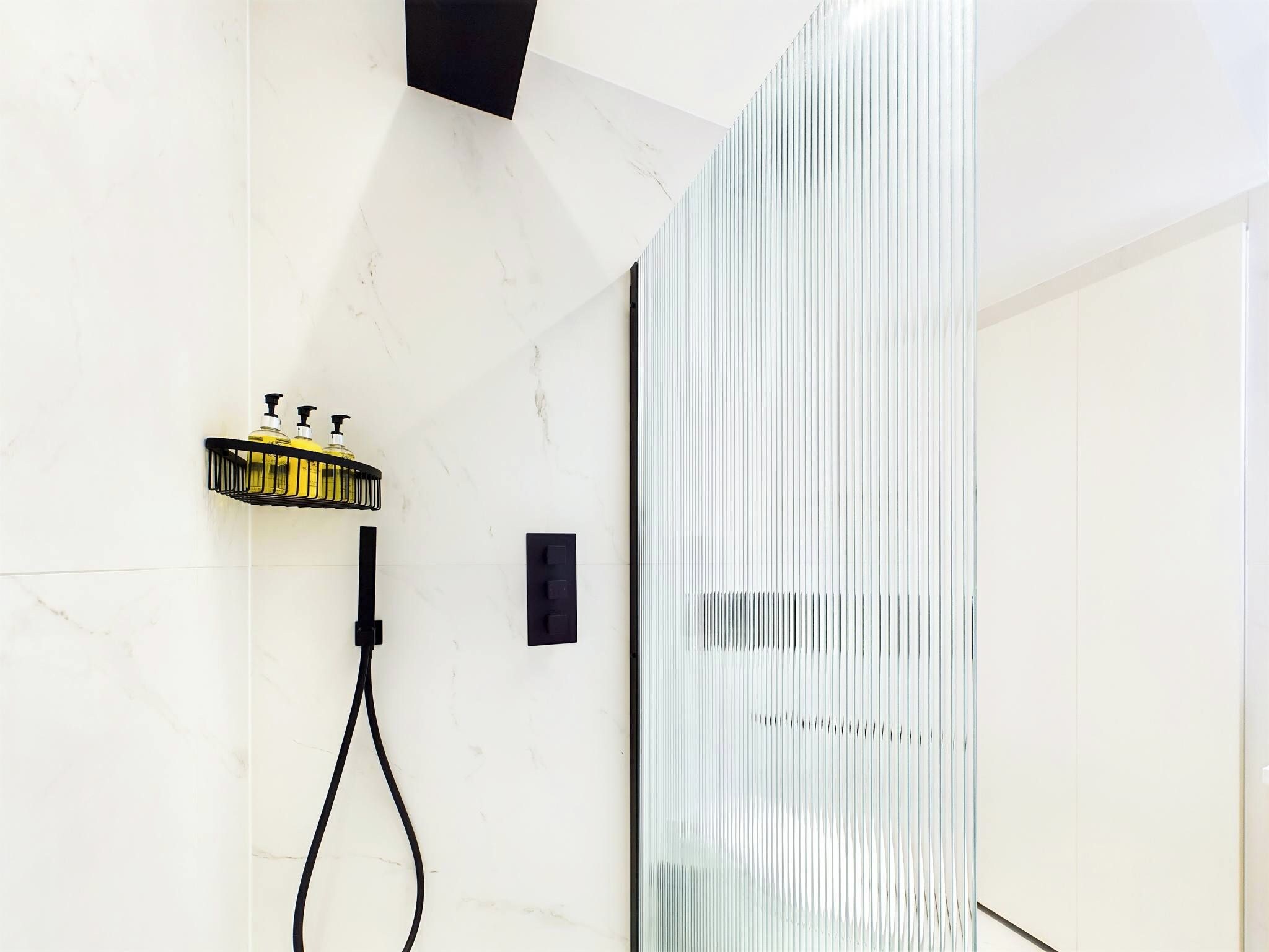 A modern bathroom with a glass shower enclosure, a black towel rack with bottles, and a marble wall.