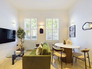 A compact living space with a green sofa, a glass coffee table, a round dining table, and a TV mounted on the wall.