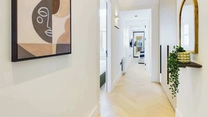 Un couloir moderne au design minimaliste, agrémenté d’un tableau abstrait aux motifs géométriques sur le mur de gauche, d’une porte blanche élégante et d’une plante posée sur une console.