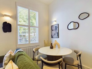 A room with a round table, four chairs, a green sofa, a window with blinds, and wall-mounted mirrors and picture frames. - Modern Maida Vale Apartment (Greater London)