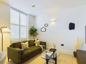 A modern living room with a green sofa, a glass coffee table, and a large window with blinds. - Decadent and Premium Apartment in Maida Vale (Greater London)