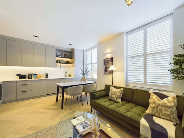 A modern living room with a green sofa, a glass coffee table, and a dining area with a white table and chairs. - Decadent and Premium Apartment in Maida Vale (Greater London)