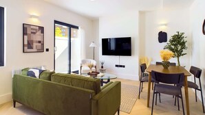 A modern living room with a green sofa, a flat-screen TV mounted on the wall, and a dining area with a wooden table and chairs.
