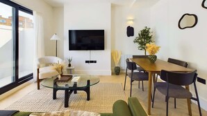 A modern living room with a glass coffee table, a sofa, a dining table with chairs, a TV mounted on the wall, and a large window.