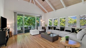 Un salon spacieux avec un grand canapé, une table basse et un téléviseur. Une porte-fenêtre coulissante donne accès à un espace extérieur avec vue sur la verdure.