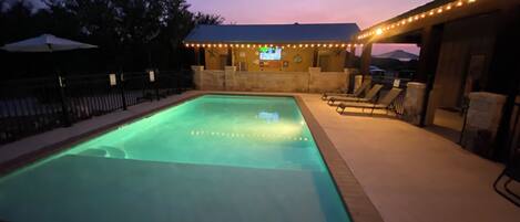 A swimming pool with a tiled deck, a covered area with seating, and a building in the background.