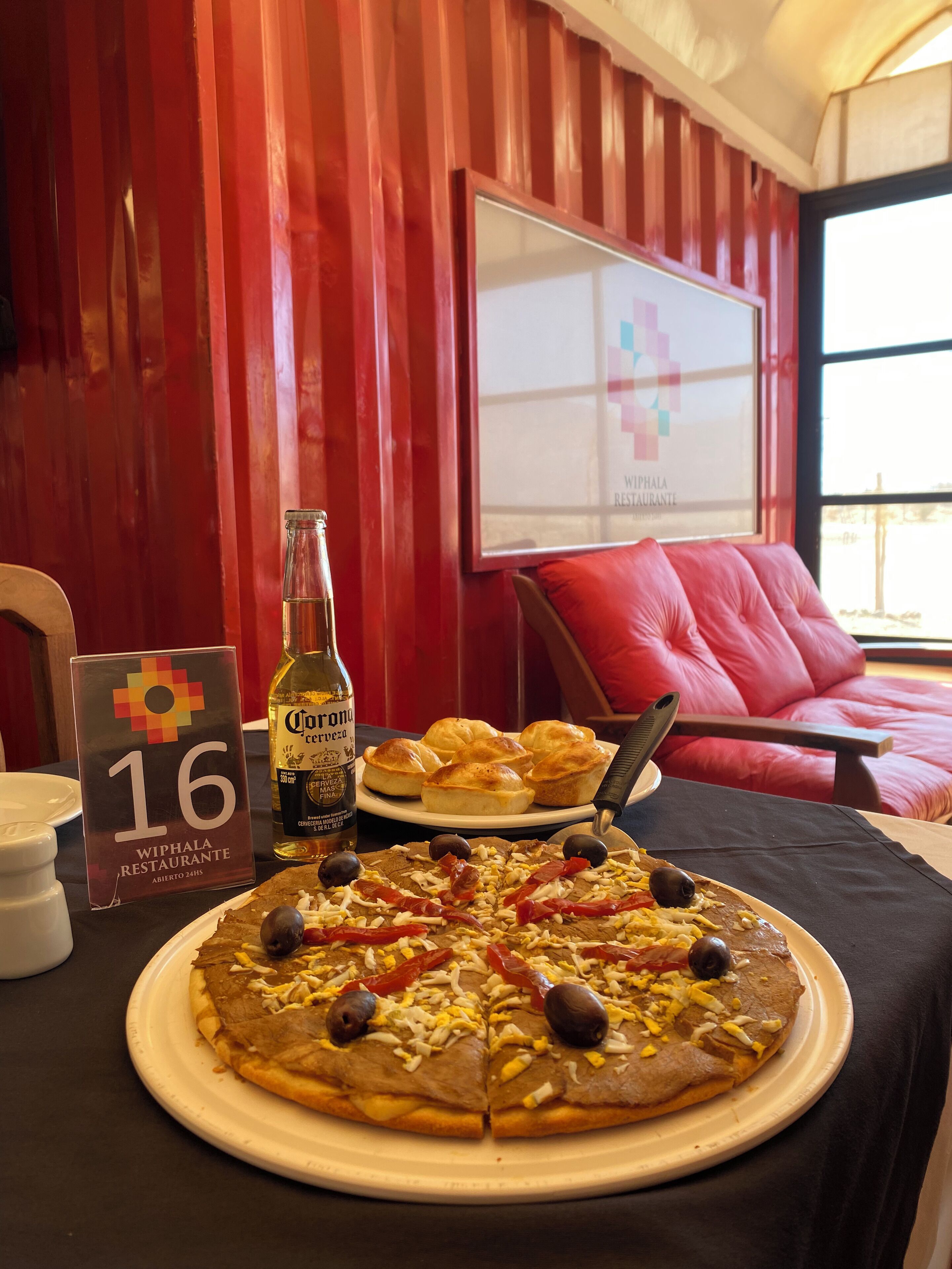Una pizza en un plat, una ampolla de Corona i un cartell de Wiphala Restaurante sobre una taula.