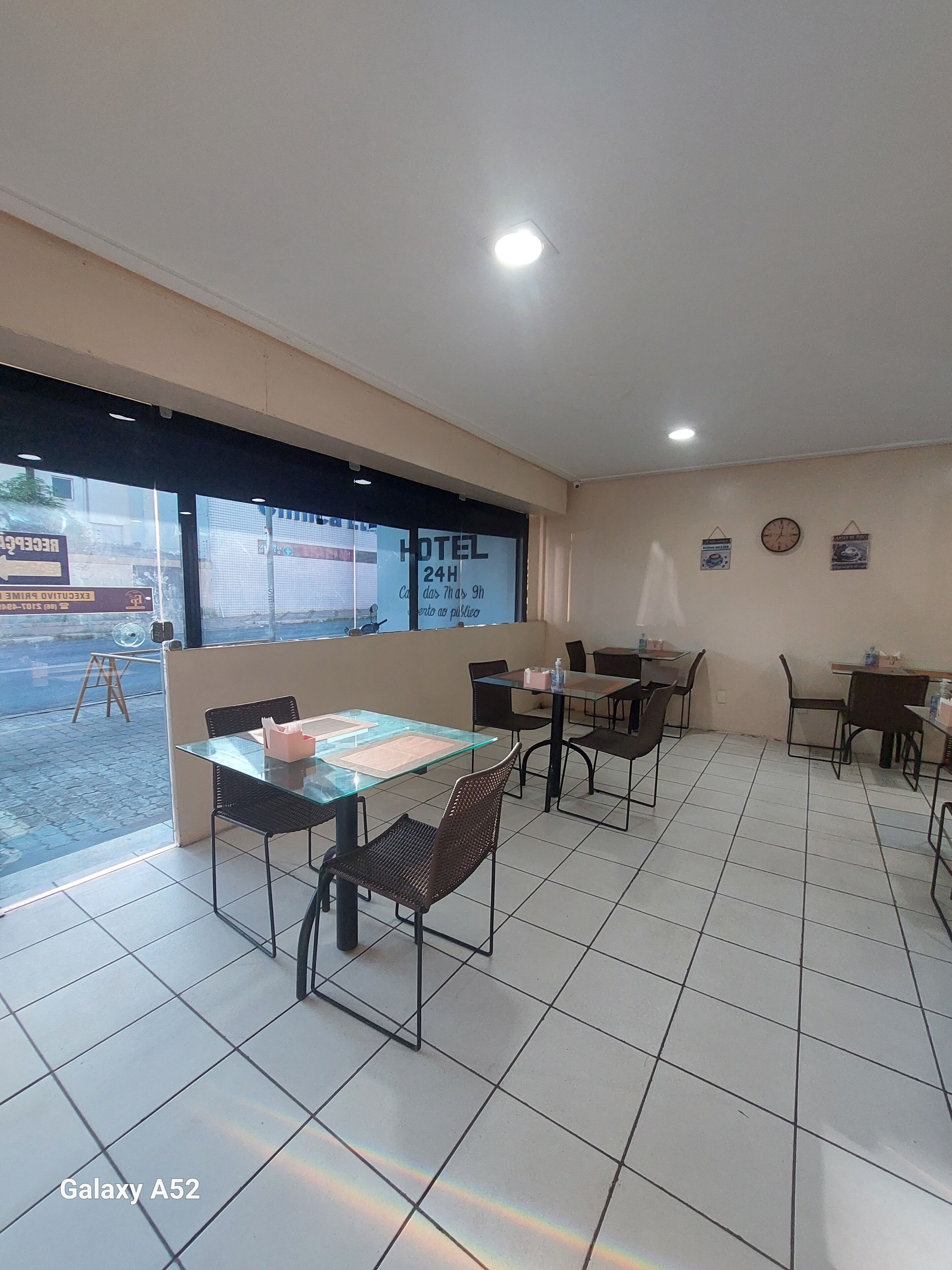 A hotel room with a tiled floor, glass tables, and chairs. There is a window with a view of the street and a sign that reads "HOTEL 24H".