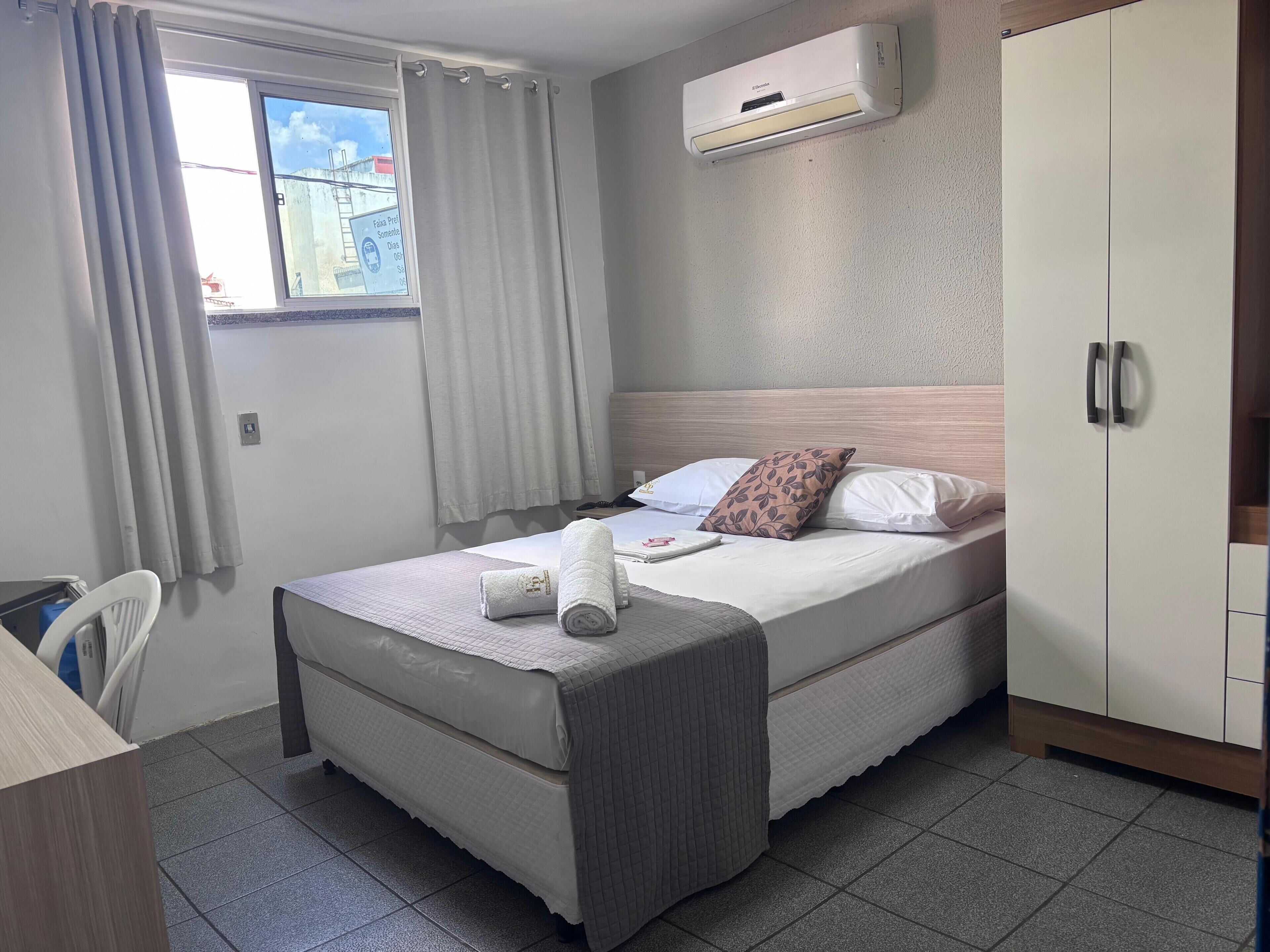 A hotel room with a bed, a wardrobe, a window with curtains, and an air conditioning unit.