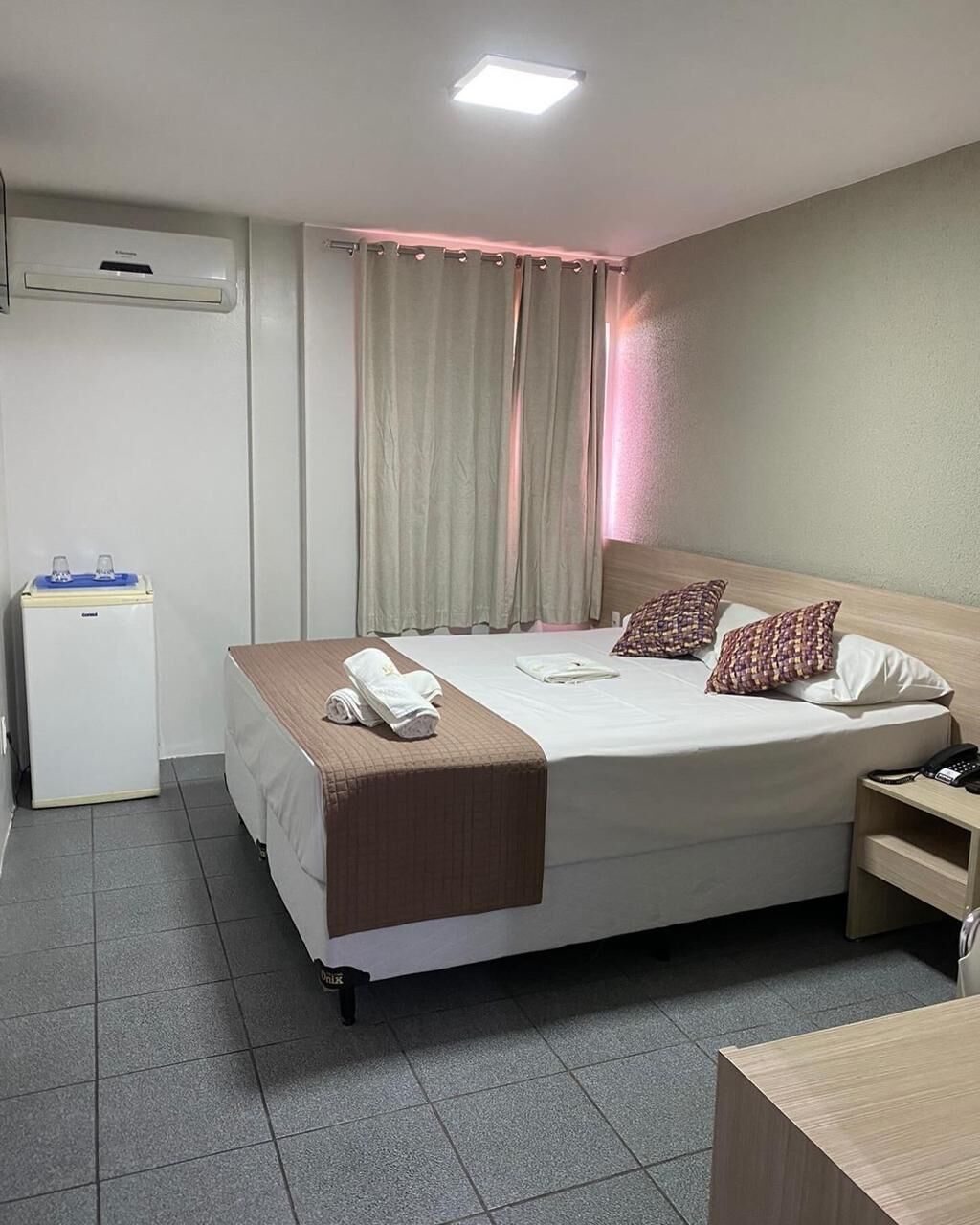 A hotel room with a bed, bedside table, lamp, and a small desk with a chair.