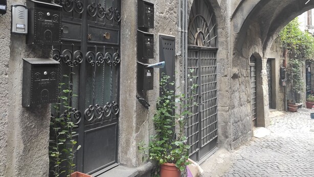Una stretta strada acciottolata con un edificio in pietra, caratterizzato da porte in ferro nero lavorate e cassette postali.