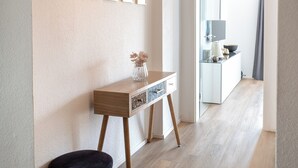 A hallway with a wooden floor, a black ottoman, a white console table with a vase, and framed pictures on the wall. - Zentral am Stadtwald (Hannover)