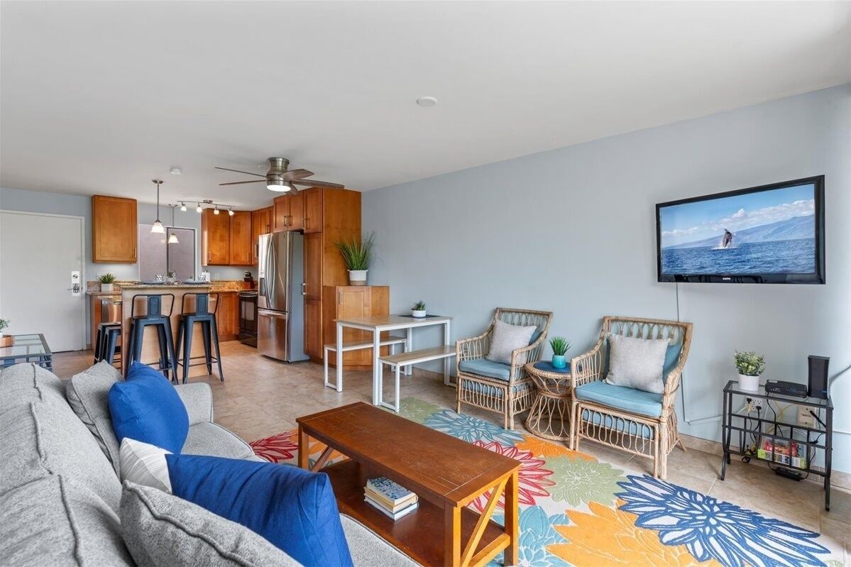 A modern living room with a sofa, coffee table, and wicker chairs. A kitchen with wooden cabinets and a bar area is visible in the background.