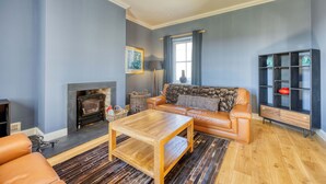 A living room with a fireplace, leather sofa, wooden coffee table, and a bookshelf. - Farmhouse Retreat with Large Hot Tub (Bishopton)