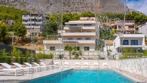 Un área de piscina con sillones de descanso y una piscina azul cristalina, rodeada de edificios con balcones y un paisaje montañoso de fondo.