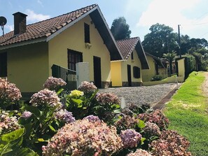 A row of yellow buildings with brown tiled roofs, a gravel path, and hydrangea flowers in the foreground. - Chalés Pedra Redonda Monte Verde Mg (Camanducaia)