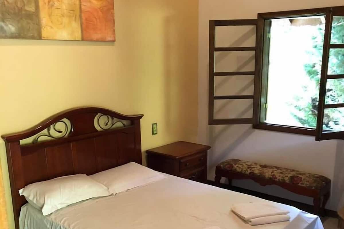 A bedroom with a wooden bed, a nightstand, a bench, and a window with a view of greenery.