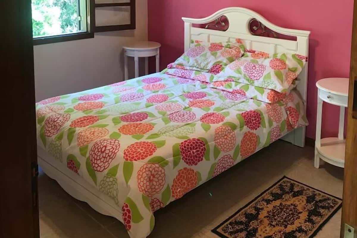 A bedroom with a bed, a window, a bedside table, a nightstand, and a painting on the wall.