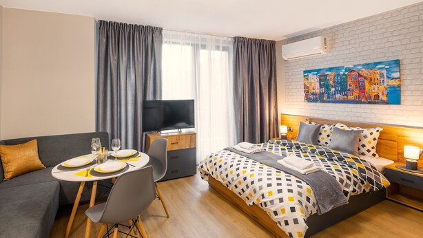 A modern hotel room with a bed, a TV, a dining table with chairs, and a painting on the wall. - Best Guest 2 Apartments (Plovdiv)