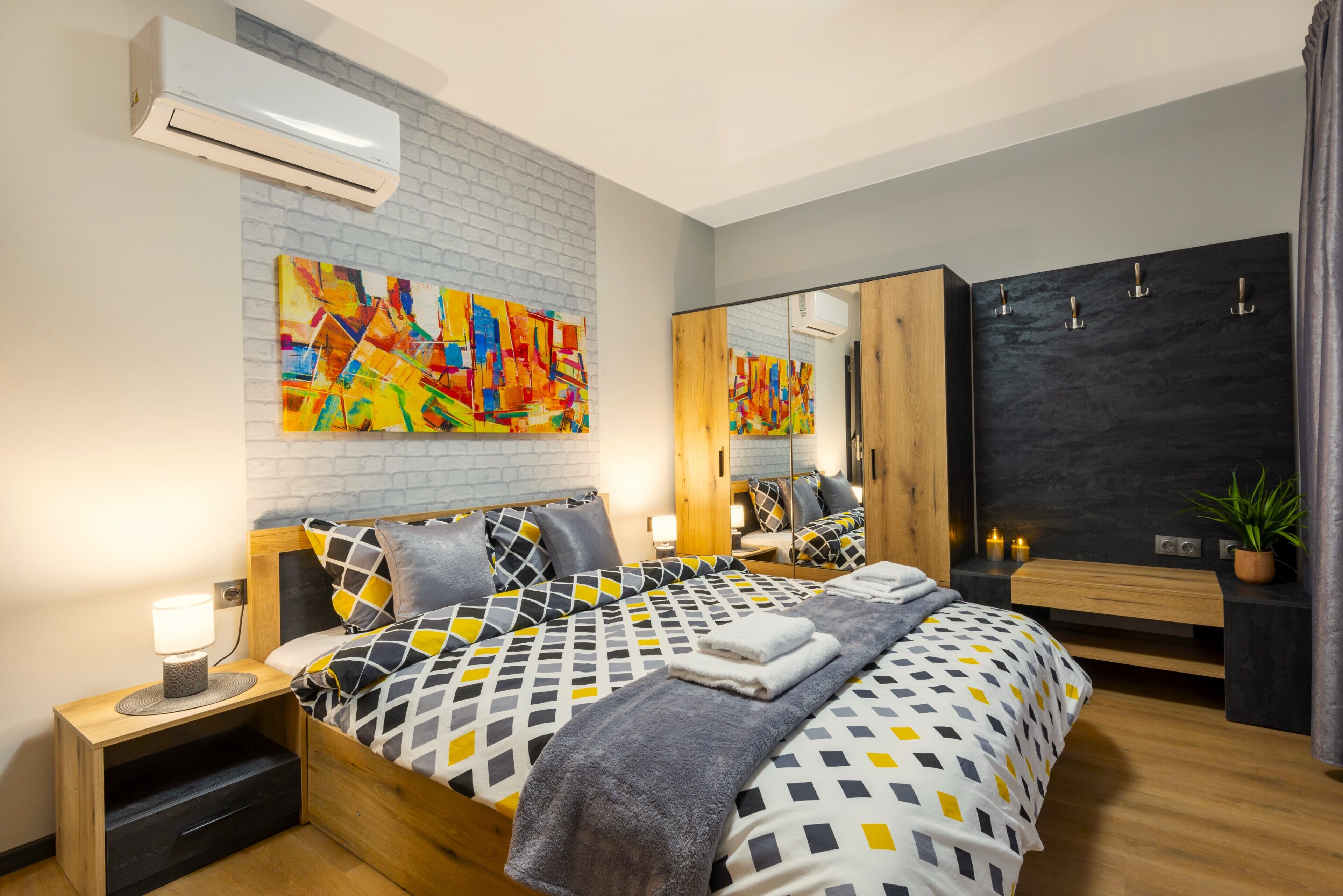 Un dormitorio moderno con una cama grande, una mesita de noche de madera, un espejo, una pizarra y un cuadro abstracto de colores.