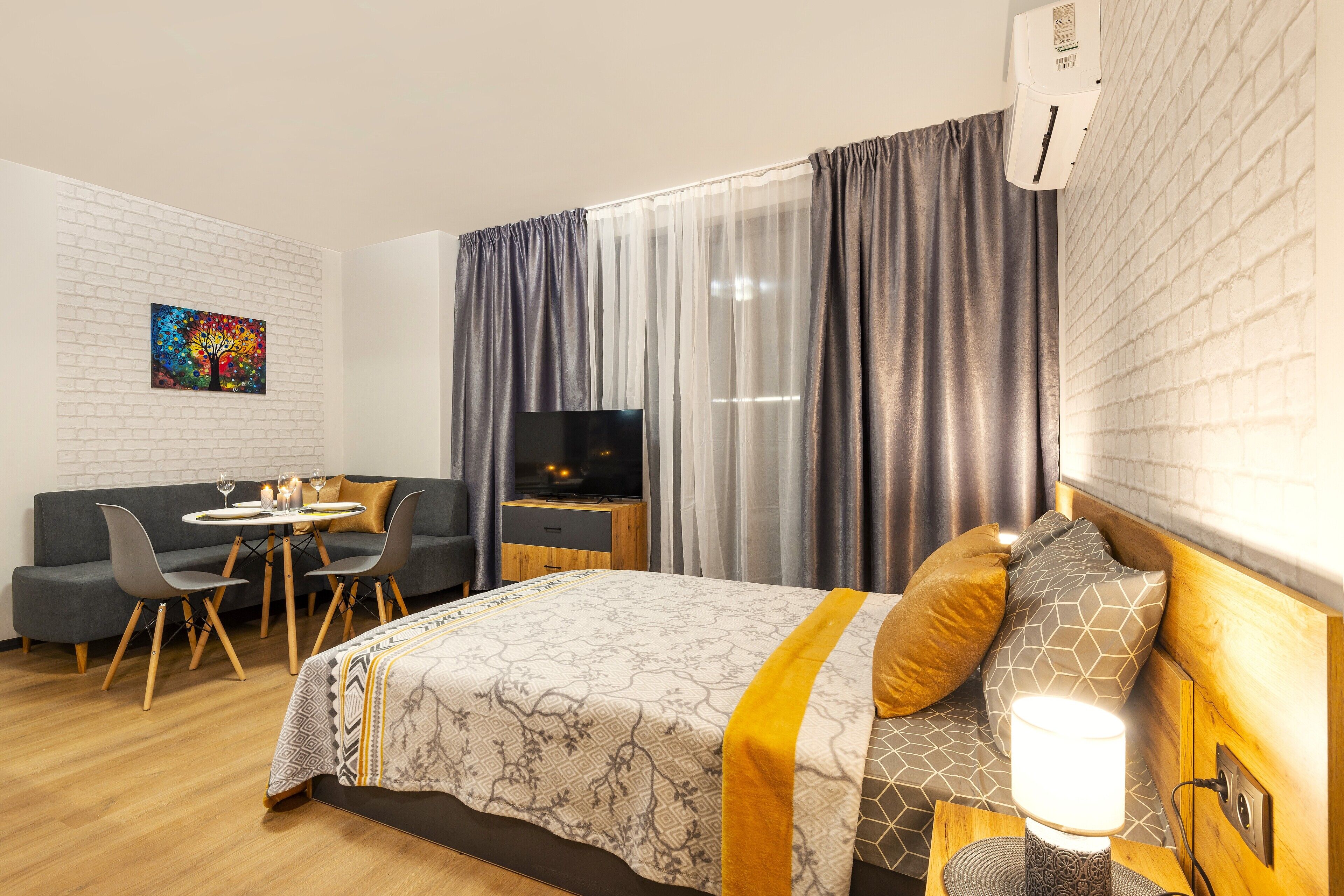 Une chambre d’hôtel moderne équipée d’un lit, d’une télévision, d’un coin repas avec une table et des chaises, et d’une grande fenêtre avec des rideaux.