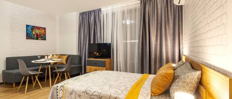 Une chambre d’hôtel moderne équipée d’un lit, d’une télévision, d’un coin repas avec une table et des chaises, et d’une grande fenêtre avec des rideaux.