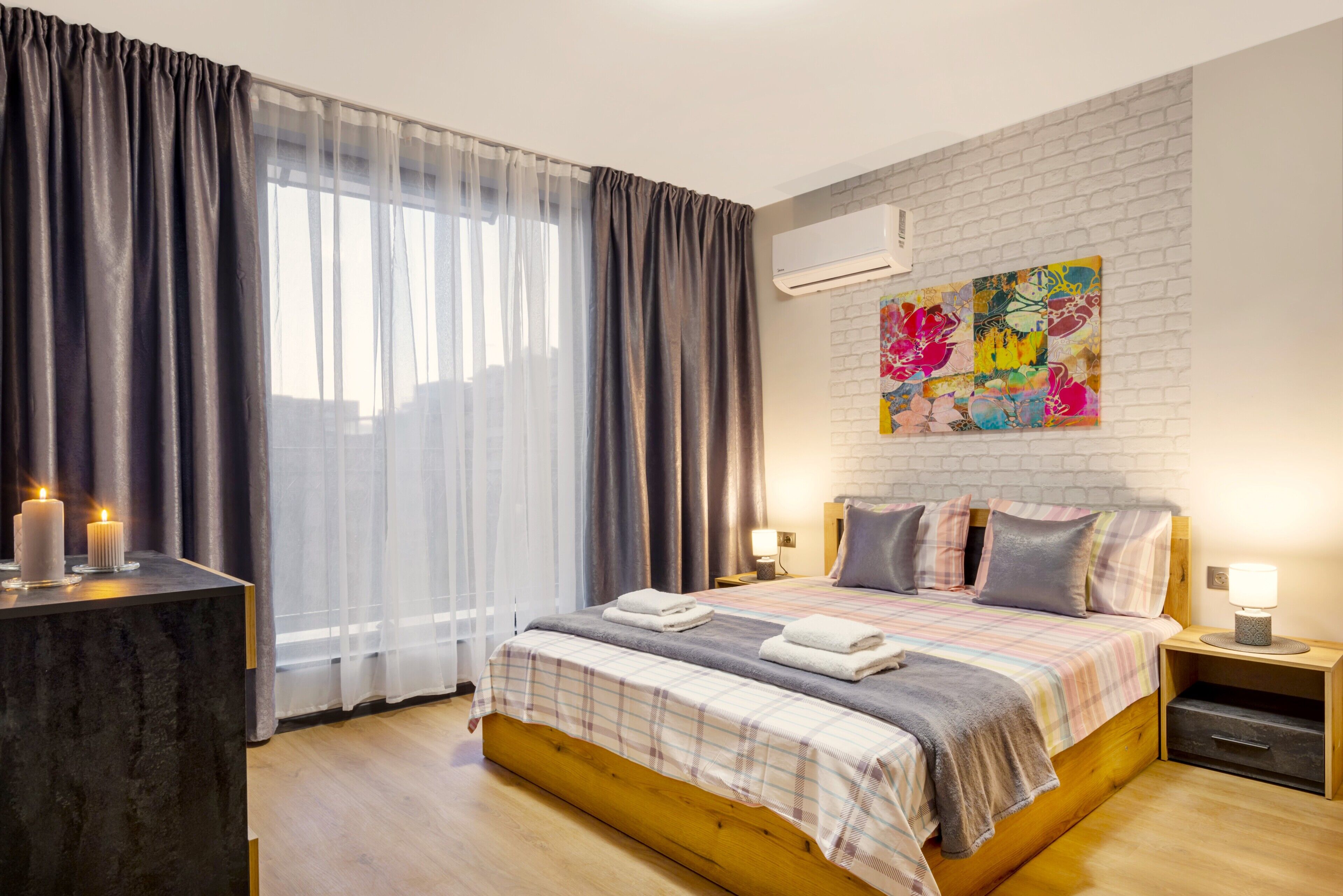 A bedroom with a wooden bed, a nightstand with a lamp, a bedside table with candles, and a colorful abstract painting on the wall.