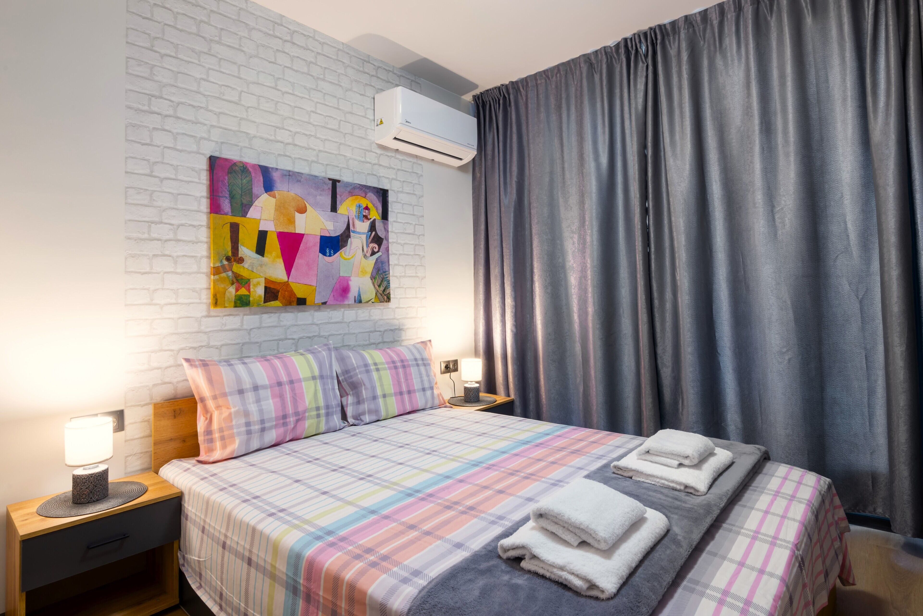 A neatly made bed with a plaid bedspread, two pillows, and folded towels on a white brick wall with an air conditioning unit above.