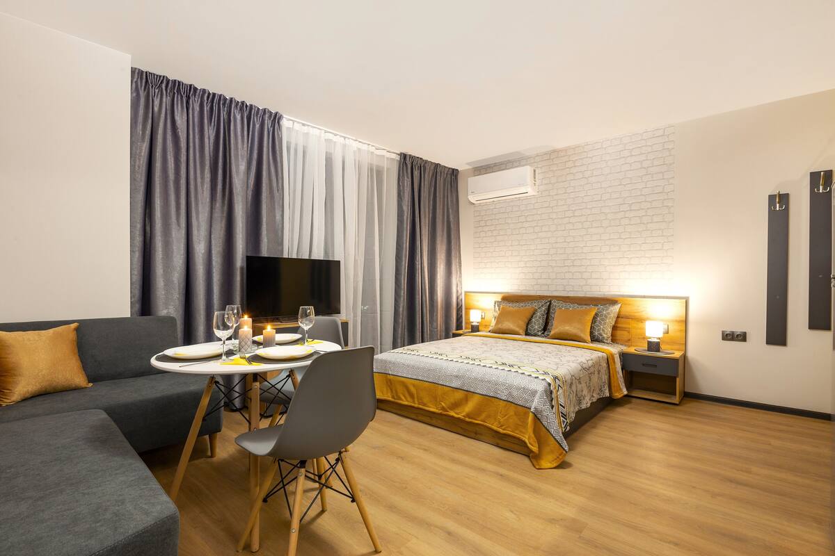 A modern hotel room with a sofa, a dining table with chairs, a bed with a headboard, and a wall-mounted air conditioner.