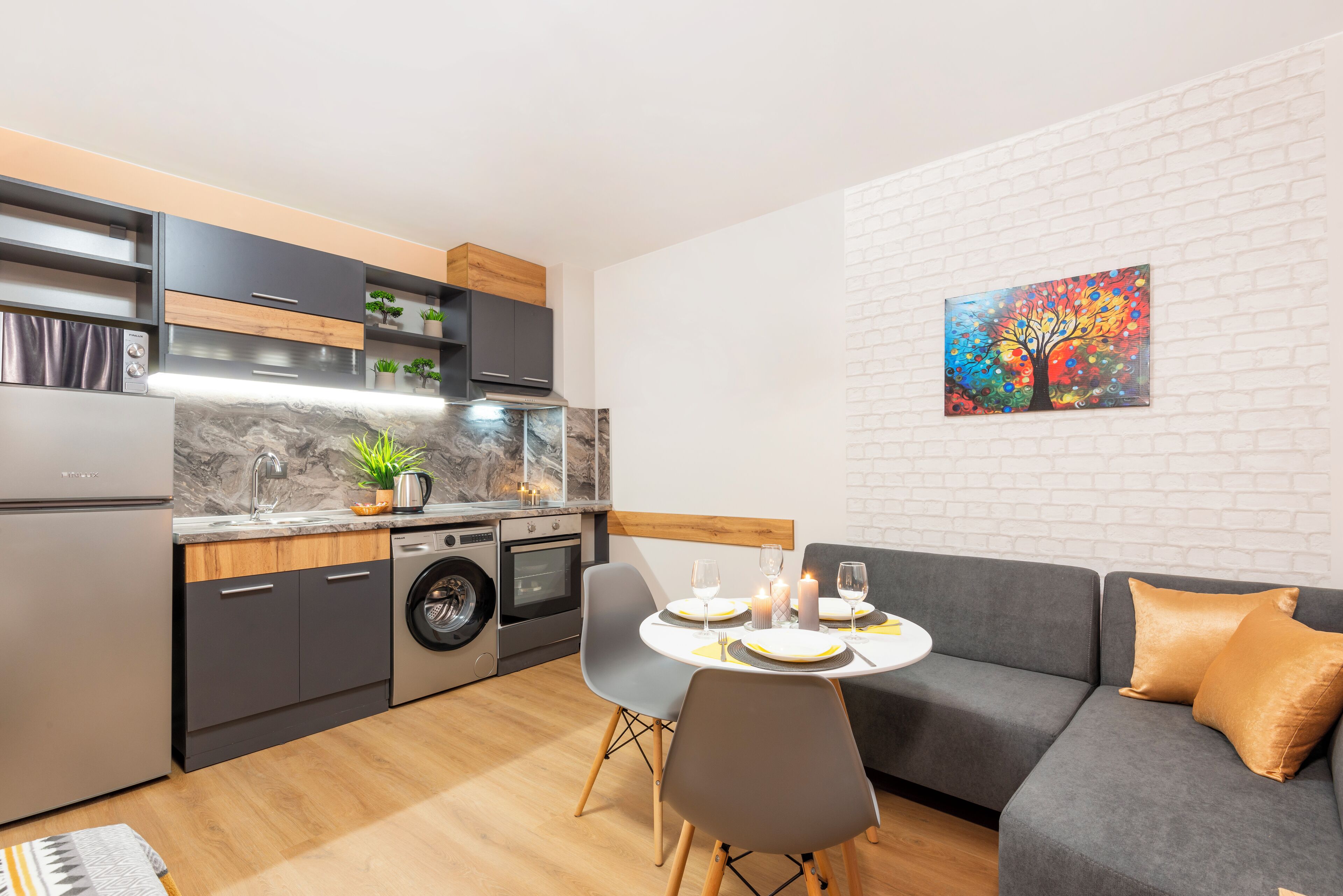 A modern kitchen with a dining area, a gray sofa, and a colorful painting on the wall.