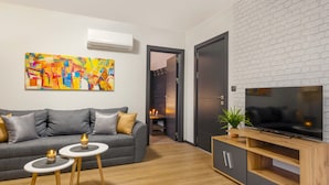 A modern living room with a grey sofa, two small tables with candles, a flat-screen TV on a wooden stand, and a colorful abstract painting on the wall. - Best Guest 2 Apartments (Plovdiv)