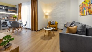 A modern living room with a grey sofa, a small round table, and a yellow armchair. There is a dining area with a table and chairs, and a kitchen area with a washing machine and a plant. - Best Guest 2 Apartments (Plovdiv)