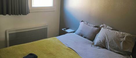 A hotel room with a bed, a window with blinds, a bedside table, and a wall-mounted light.