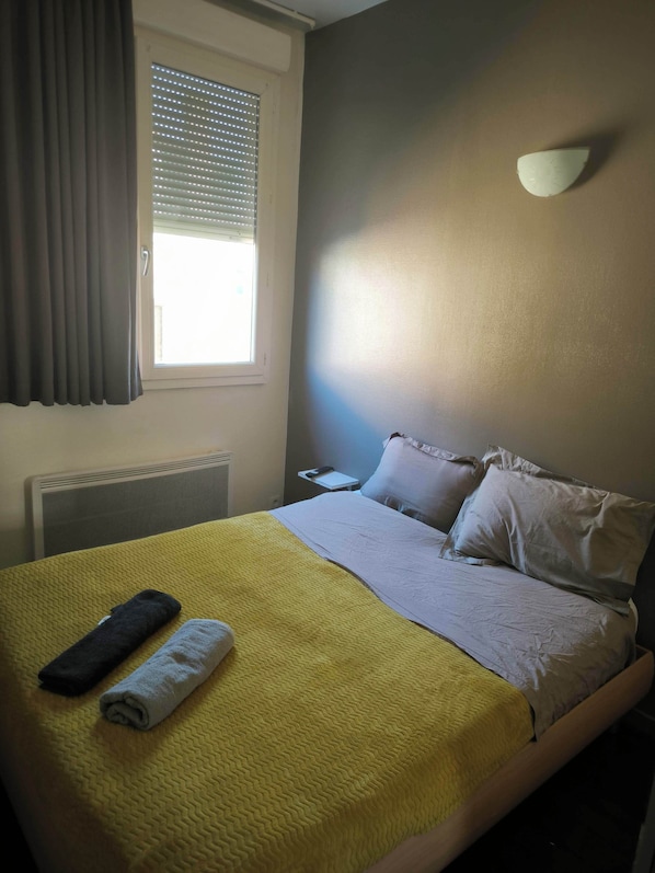 A hotel room with a bed, a window with blinds, a bedside table, and a wall-mounted light. - Hôtel de la Gare (Périgueux)