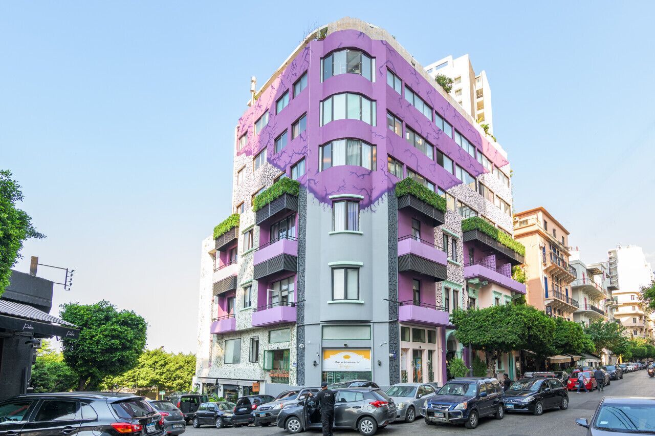 Un immeuble à plusieurs étages doté de balcons, avec des voitures garées et un ciel dégagé.