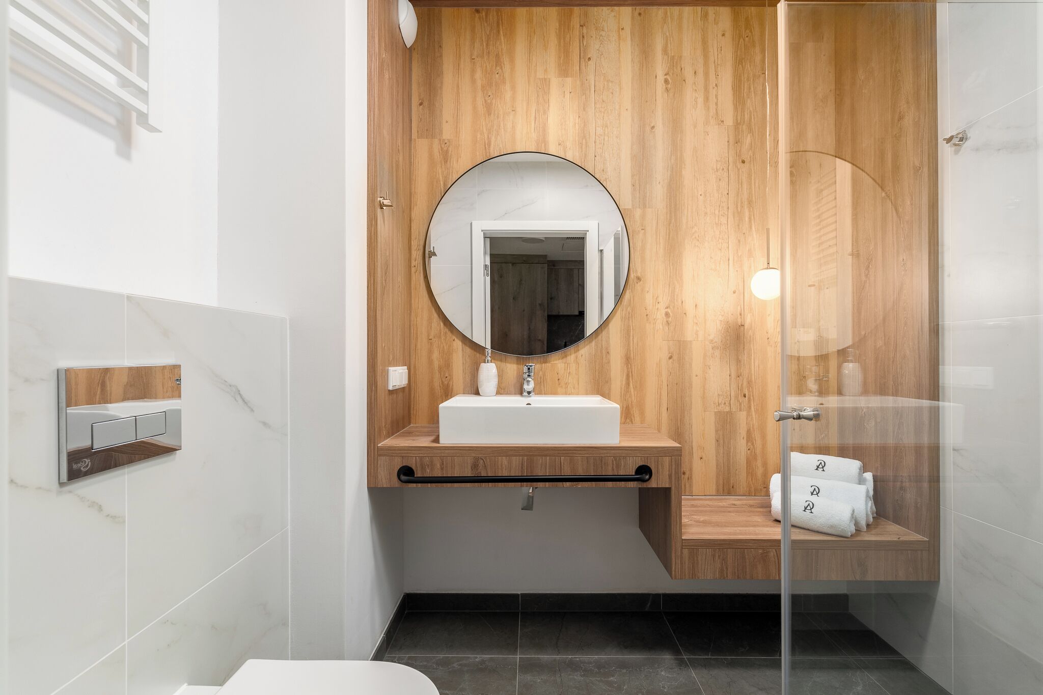 A modern bathroom with a wooden vanity, a round mirror, a white sink, and a glass shower enclosure.