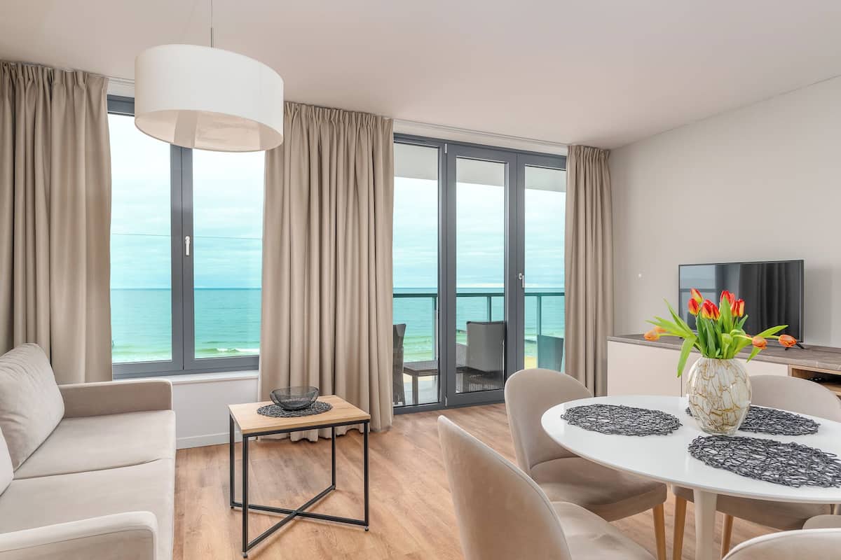 Ein modernes Wohnzimmer mit Sofa, einem Esstisch mit Platzsets, einer Blumenvase, einem Fernseher und einem Balkon mit Meerblick.