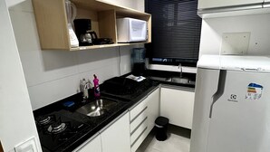 A compact kitchen with white cabinets, a black countertop, a microwave, a refrigerator, and a window with blinds.