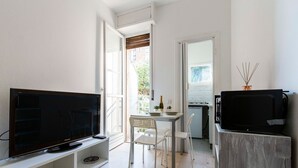 Um espaço de estar compacto com televisão de tela plana em suporte branco, uma pequena mesa de jantar com cadeiras e uma área de cozinha compacta.