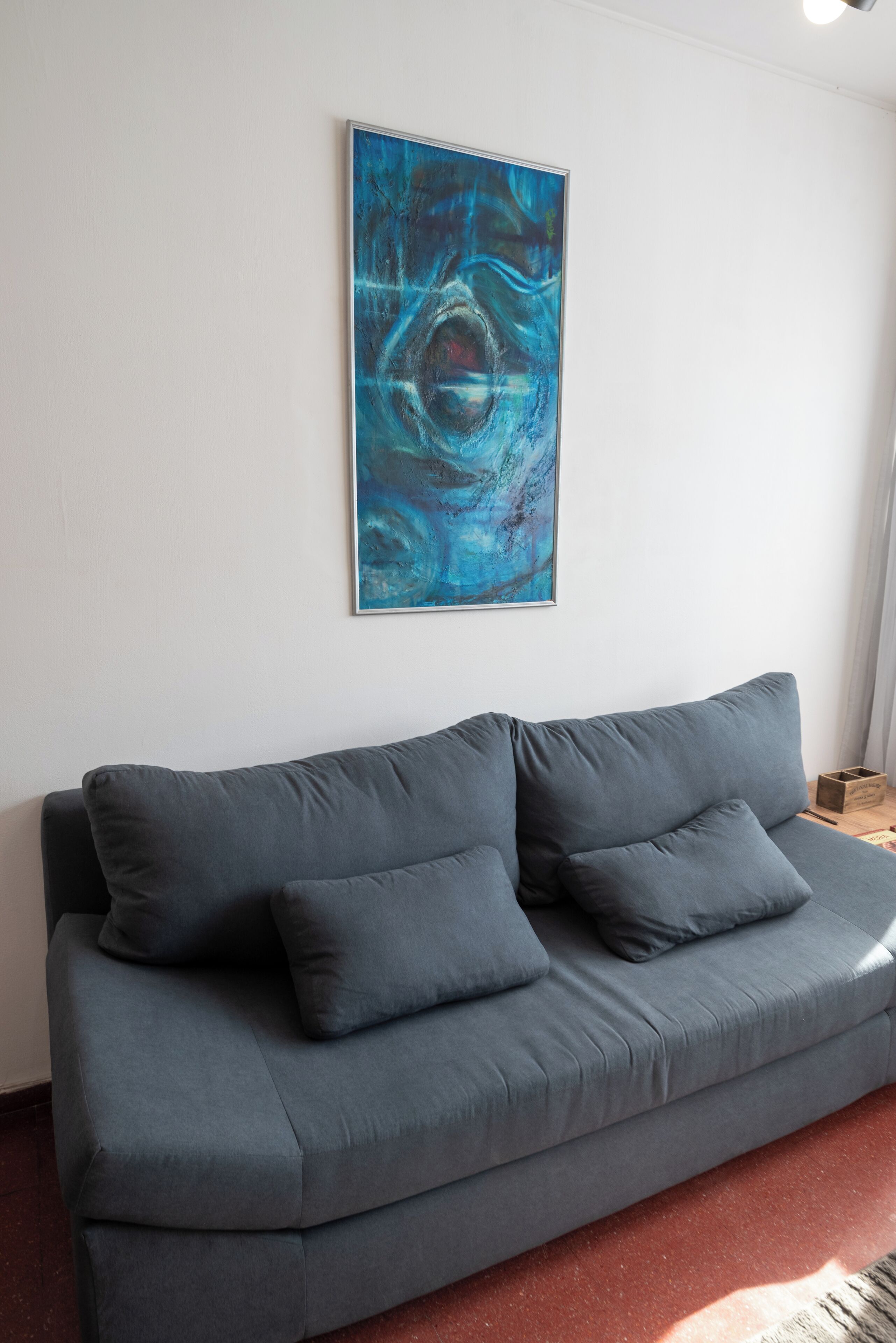 A grey sofa with three cushions against a white wall, with a blue abstract painting above it.